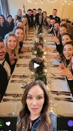 a group of people sitting at a long table with wine glasses on it and one woman standing in front of the camera