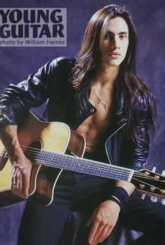 a man with long hair holding an acoustic guitar in his right hand and posing for the camera