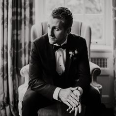 a man in a tuxedo sitting next to a window with curtains on it