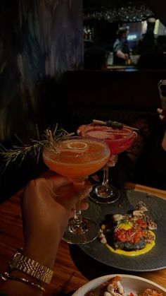 two people holding up wine glasses with food and drinks on the table in front of them