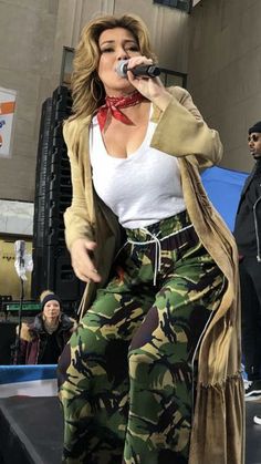 a woman standing on top of a stage with a microphone in her hand and wearing camo pants