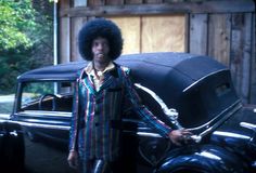 a man with an afro standing next to a car in front of a wooden building