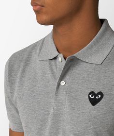 Gray Knit Polo Shirt by CdG Play, featuring a ribbed neck and cuffs. The model sports a button closure and stands out for the black heart logo patch embroidered on the chest. Casual Short Sleeve Polo Shirt With Logo Patch, Casual Cotton Polo Shirt With Logo Patch, Casual Polo Shirt With Logo Patch, Casual Polo Collar Top With Embroidered Logo, Collared Cotton Top With Logo Detail, Designer Cotton Tops With Ribbed Collar, Casual Polo Shirt With Embroidered Logo For Streetwear, Casual Cotton Polo Shirt With Logo Detail, Casual Cotton Polo Shirt With Logo