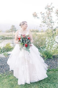 a woman in a white wedding dress holding a red and green bouquet standing on the grass