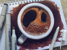 a cup of coffee on top of a spiral notebook next to markers and pencils