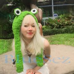 a woman wearing a green frog hat with big eyes sitting on a bench in front of a statue