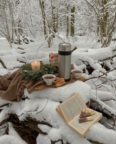 an open book sitting on top of snow covered ground next to a cup and candle