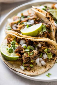 three shredded pork tacos on a white plate with limes and cilantro