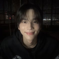 a young man smiling at the camera in front of a window with city lights behind him