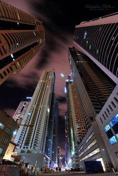 some very tall buildings at night time with lights on and clouds in the sky above them