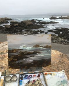 an artist's easel with paint and watercolors in front of the ocean