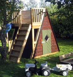 a small wooden play house with a slide and ladder to the top, next to a lawn mower