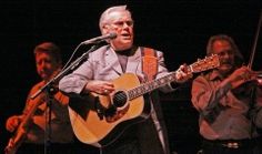 an older man playing the guitar and singing into a microphone while other men stand behind him