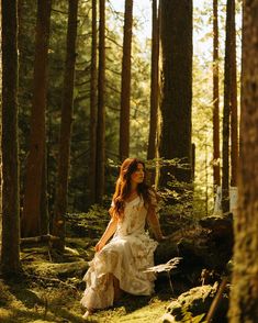 a woman in a white dress is sitting on the mossy ground surrounded by trees