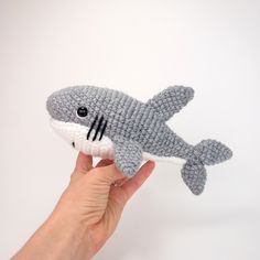 a hand holding a knitted stuffed shark toy in front of a white wall background