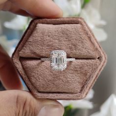 a person holding an engagement ring in a box with white flowers behind it on a table