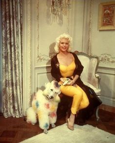 a woman sitting on a chair next to a white poodle in front of a chandelier