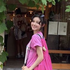 a woman in pink carrying a black handbag and smiling at the camera while standing next to a tree