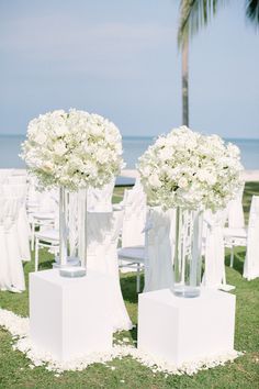 two tall vases with white flowers are on the grass near chairs and palm trees