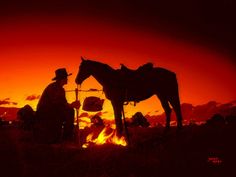 two people standing next to a horse in front of a fire with the sky red and orange