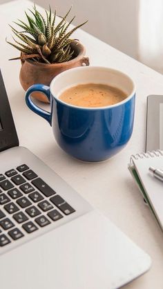 a cup of coffee sitting next to a laptop computer on a white desk with a plant