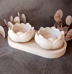 two white vases sitting on top of a table next to some leaves and flowers