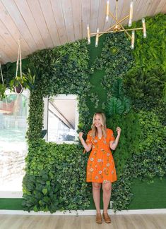 a woman in an orange dress standing next to a green wall with plants on it