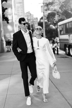 a man and woman are walking down the street in front of a bus wearing sunglasses