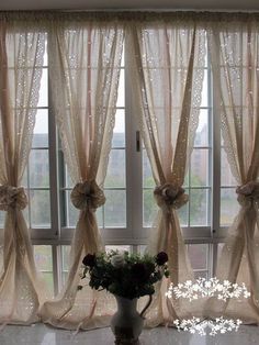 a vase filled with flowers sitting next to two windows covered in sheer curtaines and bowknots