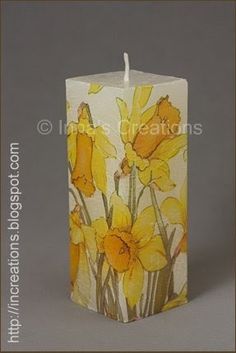 a candle with yellow flowers painted on the front and sides, sitting next to a gray background