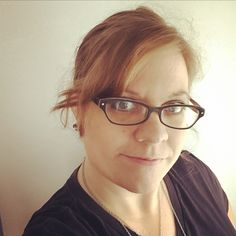 a woman with glasses and a necklace on her neck looking at the camera while standing in front of a white wall