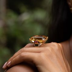 The Story Bright and bold, this vintage cocktail ring from the Retro era is a gorgeous piece. Done in warm 14 karat yellow gold, the ring has an allover glow. At center is an extra-large emerald cut citrine weighing approximately 55 carats. The hand-cut stone sits in a four-prong setting made for the piece. On either side are four ruby cabochons that look like juicy pomegranates seeds. Moving down the band are six vintage single cut diamonds set in 14k white gold giving a sparkle filled finishin Citrine Cocktail Ring, Yellow Gemstone Jewelry, Smokey Topaz Ring, Cocktail Ring Designs, Retro Era, Yellow Style, Vintage Cocktail Ring, Jewelry Staples, Smokey Topaz