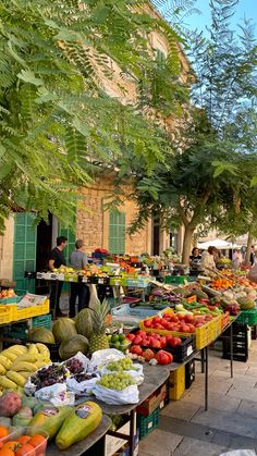 an outdoor market with lots of fruits and vegetables on tables in front of trees, people shopping