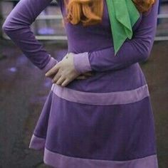 a woman in a purple dress and green hat is standing on the street with her hands on her hips