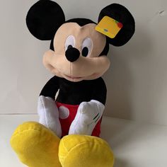 a mickey mouse stuffed animal with a yellow tag on its ear and red shorts, sitting in front of a white wall