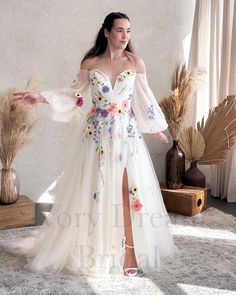 a woman wearing a white dress with flowers on the side and long sleeves, standing in front of a window