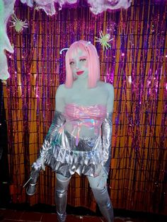 a woman with pink hair and silver outfit standing in front of a wall covered in lights