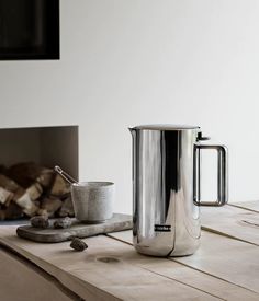 a stainless steel coffee pot sitting on top of a wooden table next to a fire place