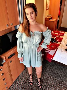 a woman standing in front of a bed wearing sandals and an off the shoulder dress