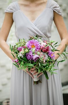 a woman in a gray dress holding a bouquet of purple and white flowers on her wedding day