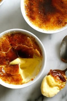 two white bowls filled with food on top of a marble counter next to spoons