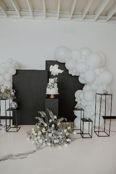 a white wedding cake surrounded by balloons and flowers on a black stand in front of a wall