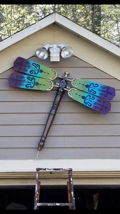 a colorful dragonfly clock mounted to the side of a house next to a ladder