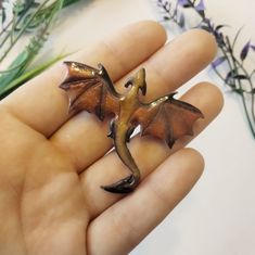 a hand holding a ring with a small dragon on it's finger and flowers in the background