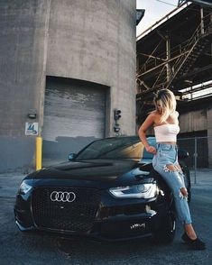 a woman leaning on the hood of a black car in front of an industrial building