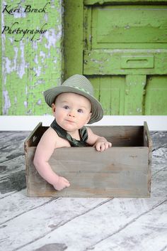 a baby wearing a hat and tie sitting in a wooden box on the floor next to a green door