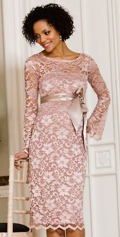 a woman in a pink lace dress standing next to a chair and smiling at the camera