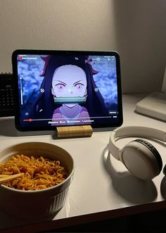 a bowl of noodles and headphones on a desk next to a computer monitor with an anime character