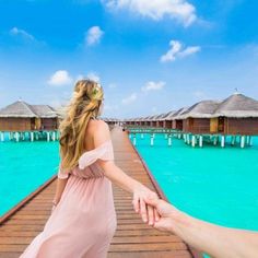 two people holding hands while walking across a wooden bridge over water with thatched huts in the background