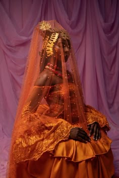 a woman in an orange dress and veil sitting on a purple surface with pink curtains behind her